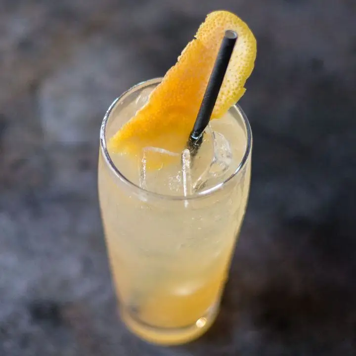 A top-down shot of a golden-hued drink filling a highball glass, with a black paper straw and a long grapefruit peel garnish. The background is charcoal colored. 