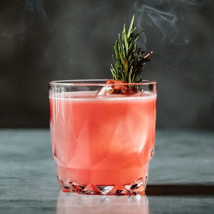 A faceted rocks glass rests on a gray marble surface. The glass is filled with a light red drink, some ice cubes and a gently smoking sprig of rosemary.