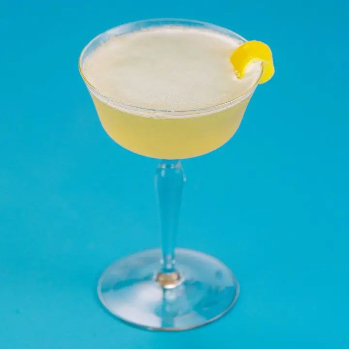 An elegant, thick-stemmed cocktail glass is filled with a hazy yellow drink and garnished with a lemon peel draped over its lip. The background is solid blue.