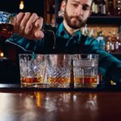 A bearded bartender in a blue plaid shirt pours a stream of Irish whiskey into a patterned rocks glass. Two other glasses flank the center one that he is pouring into, each with a large dram of whiskey.