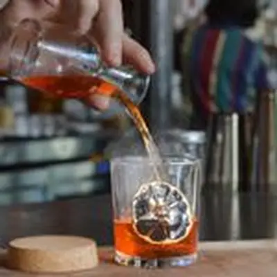A bartender pouring a ruby-red cocktail into a rocks glass containing a dehydrated citrus wheel