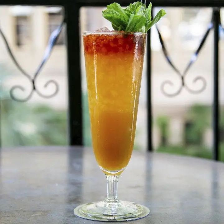 A tall glass holds a multi-layered drink, going from deep red at the top to orange mixed with crushed ice and then a thick and opaque orange base. The drink is garnished with a generous sprig of mint, and served on a table on a balcony.