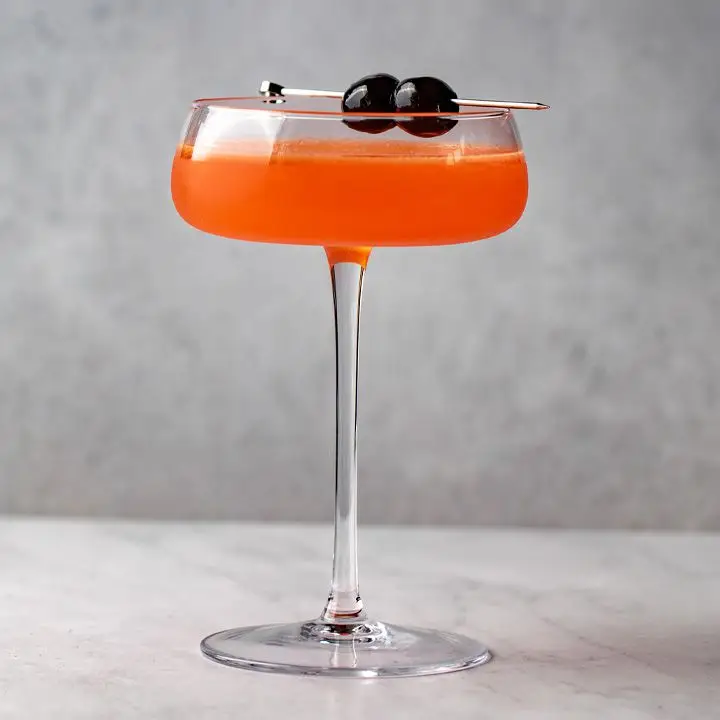 A long-stemmed coupe with a short flat bowl holds a bright orange cocktail. The drink is garnished with two dark cherries on a metal pick. The background and floor are both gray marble.