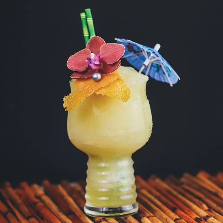 Frozen yellow Saturn cocktail in hurricane glass with orchid and orange peel garnishes, bamboo straws and cocktail umbrella