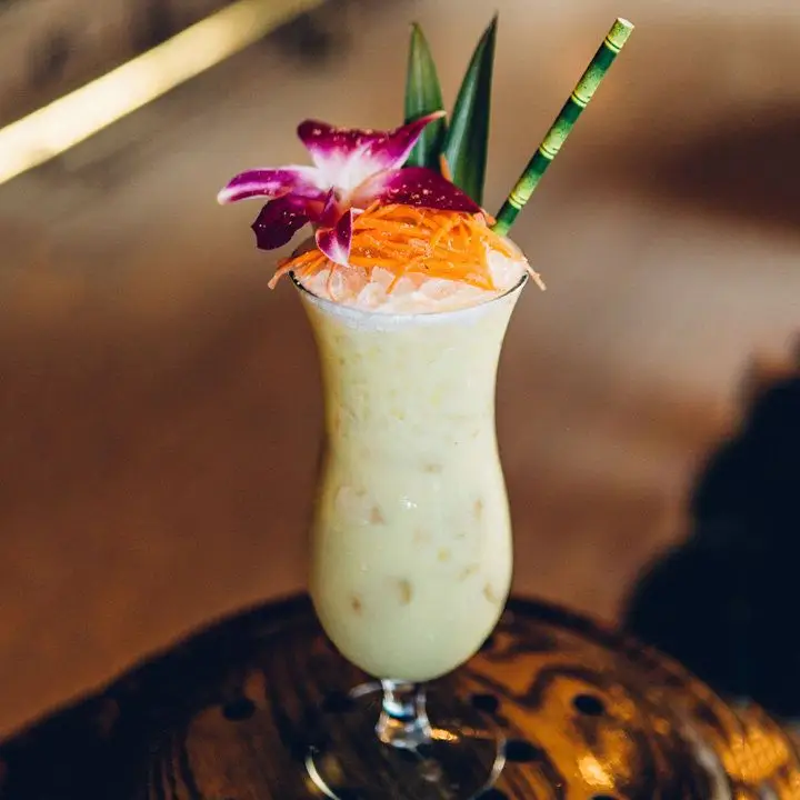Gangsters Paradise Pina Colada riff cocktail served in hurricane glass with crushed ice, topped with orchid, shaved carrots and pineapple frond with green straw 