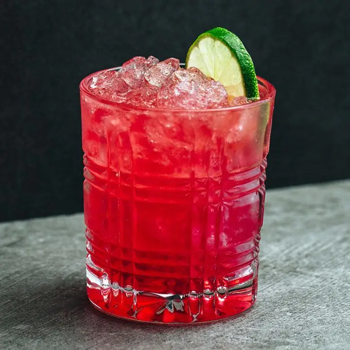 Vibrant pink Maracuya Mistress cocktail in rocks glass with crushed ice and lime wheel, shot against black background on gray surface