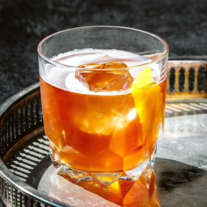 On a metal bar tray, a chunky rocks glass holds a bright orange cocktail. An ice cube pokes from the top of the drink, as does a thin slice of lemon peel.