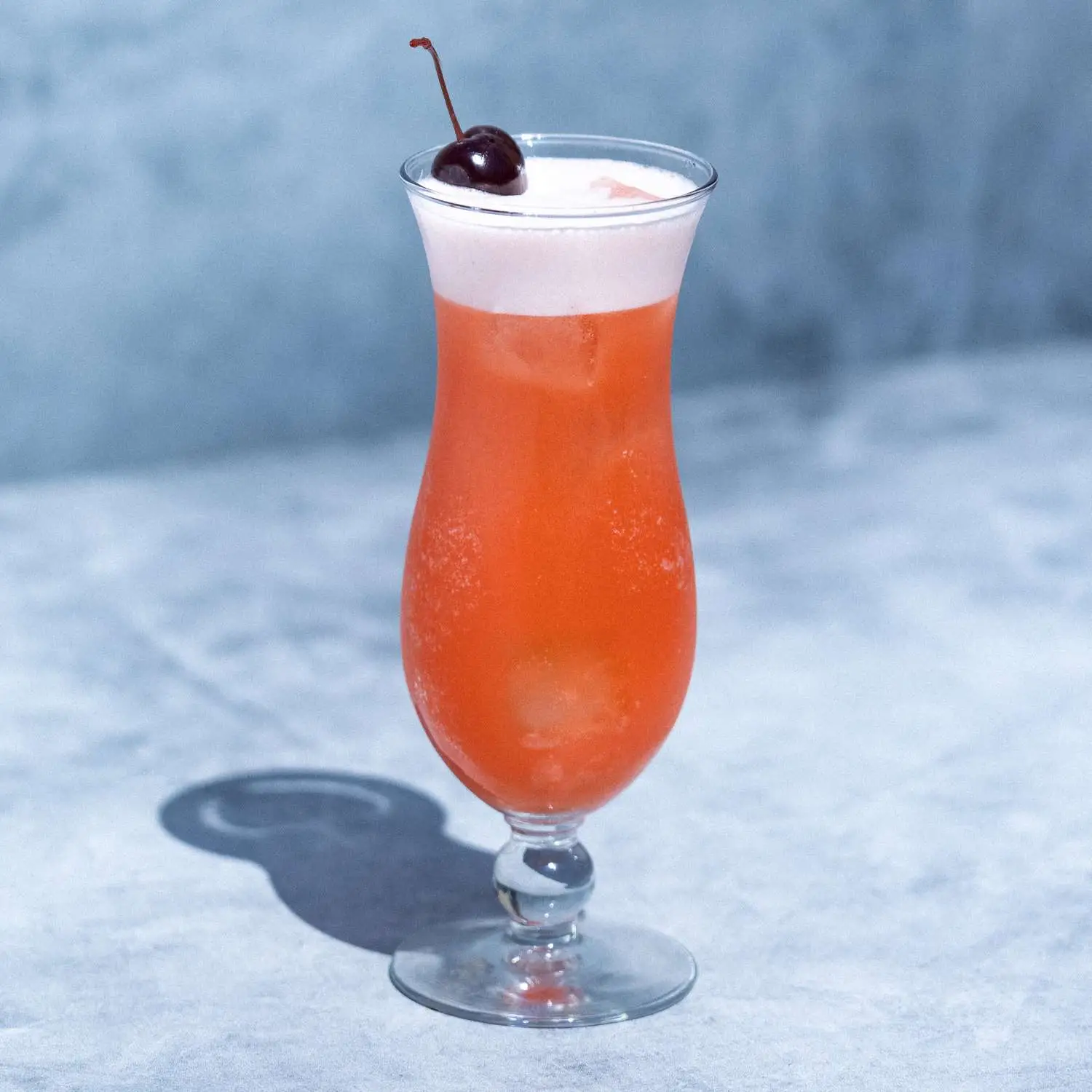Orange-red Rum Punch in a curved Hurricane glass with a thick white head on top, garnished with a single maraschino cherry