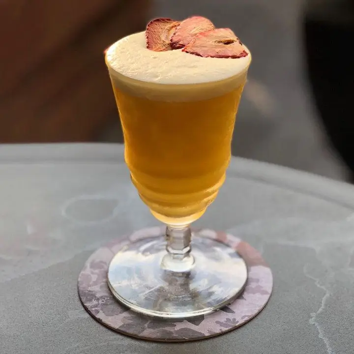 An unusual tall, knobbed cocktail glass holds an opaque orange-brown drink with thick foam. It’s garnished with a few dried strawberry slices.