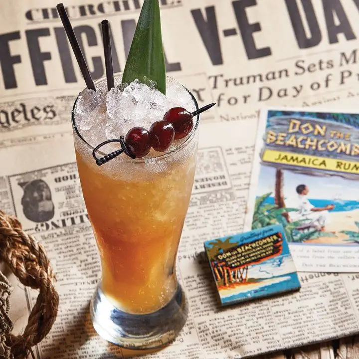 A footed pilsner glass holds a dark rum drink and crushed ice, garnished with a pineapple frond and three cherries skewered on a sword-shaped pick. The drink is placed on top of a newspaper announcing the Allies’ victory over the Axis in World War II, as well as some Tiki ads featuring Don the Beachcomber