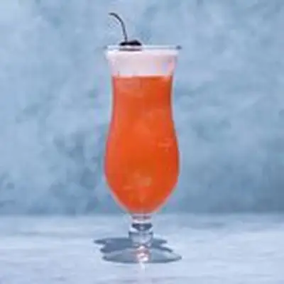 Orange-red Rum Punch in a curved Hurricane glass with a thick white head on top, garnished with a single maraschino cherry