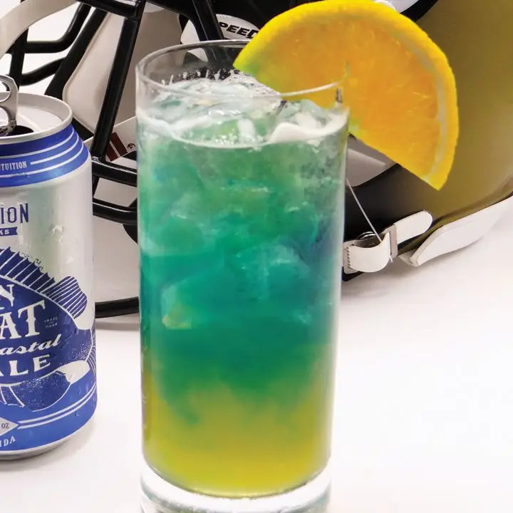 A Collins glass is filled with a golden drink with a deep blue float; the two colors slowly mix together. It’s garnished with a large slice of orange, and a football helmet is behind it.