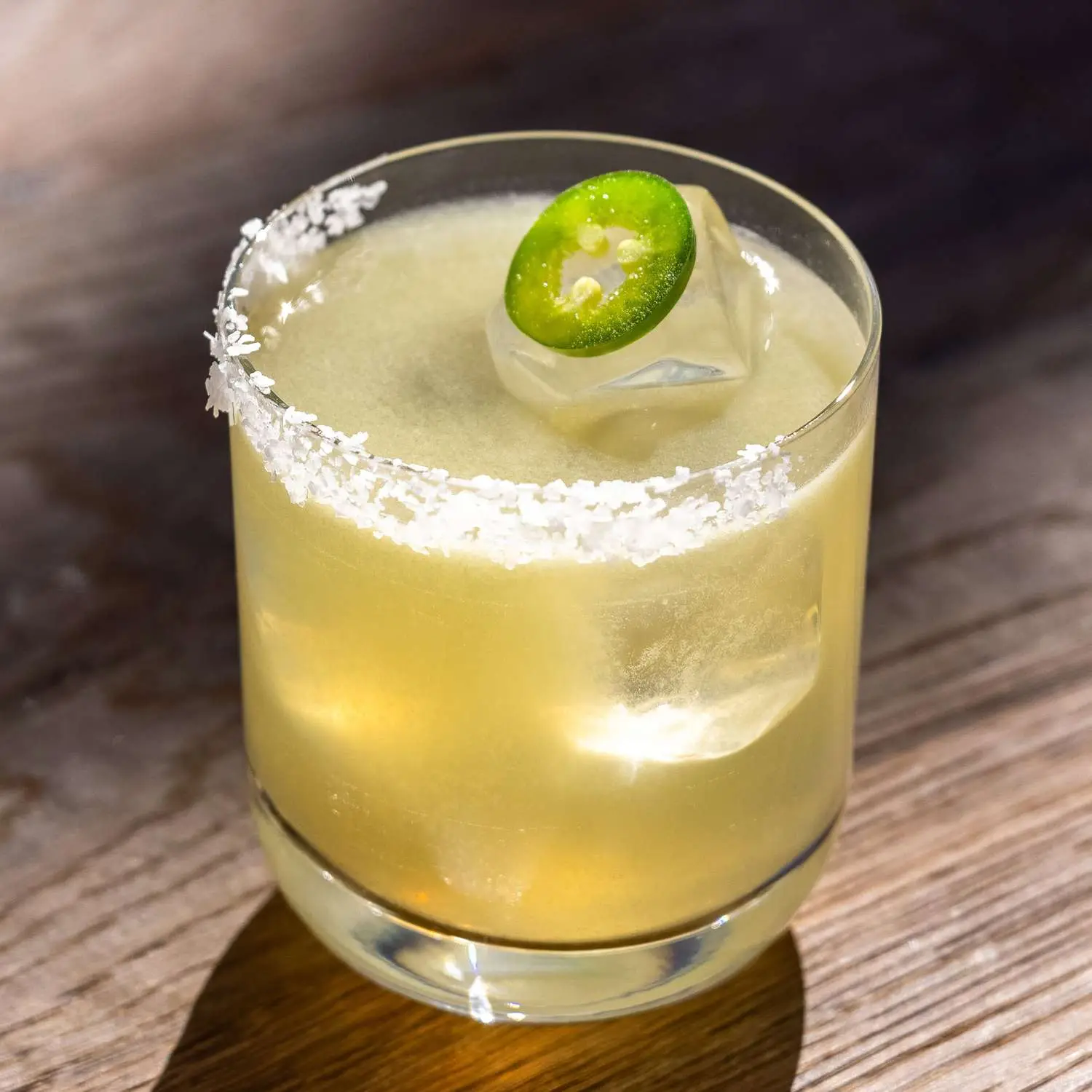Spicy Margarita in ice-filled rocks glass with half salt rim and jalapeno coin garnish, shot on wooden surface