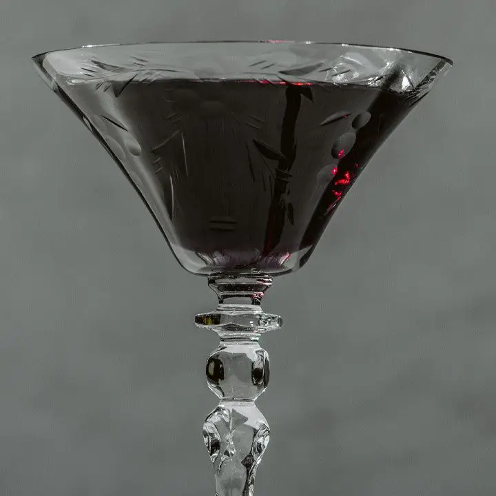 A small, elegant, knobbed cocktail glass holds a dark, crimson drink. The glass is set against a neutral gray background.