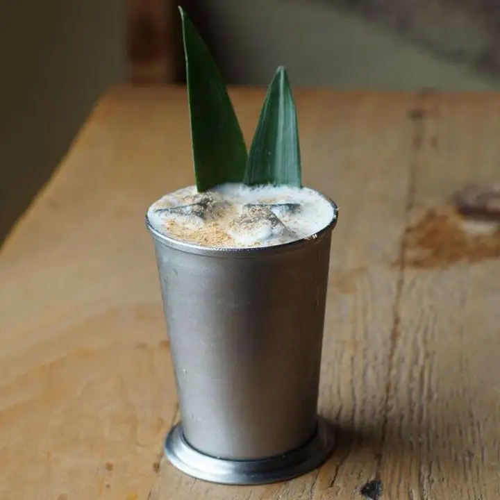 A silver Julep cup rests on a rustic wooden table. The cup is filled with a foamy white drink, dusted with nutmeg and garnished with pineapple fronds. 