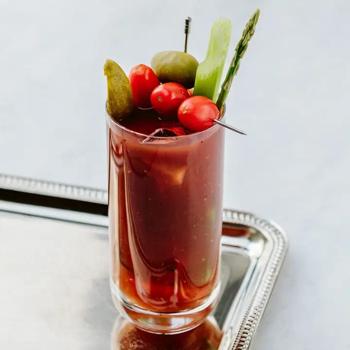 Bloody Bulldog cocktail in a highball glass, loaded with garnishes (tomatoes on a skewer, pickles, olive, asparagus), served on a silver tray