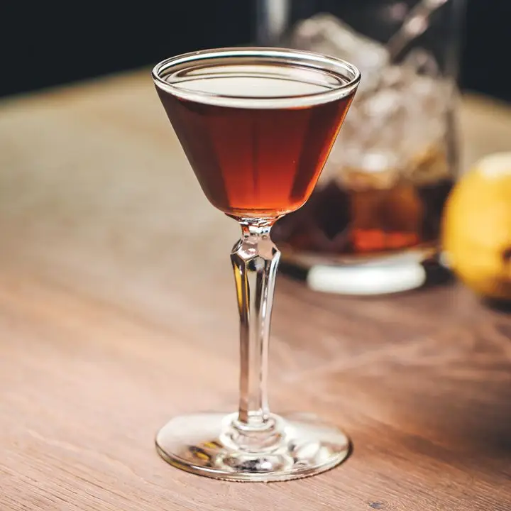 A thin cocktail glass with a knobbed stem rests on a wooden table. It’s filled with an amber drink, and behind it a mixing glass holds ice and more of the drink. 