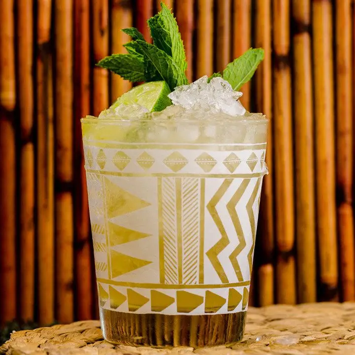A Tiki glass with white markings holds a golden Mai Tai. Crushed ice, mint and a lime wedge pick from above the lip of the glass. The background is wood lines, and the drink rests on a wicker mat.