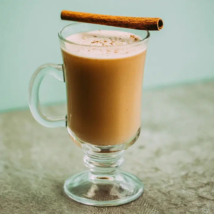 A glass mug holds a lush, golden-brown serving of Hot Buttered Rum. Its pale surface is flecked with nutmeg, and a cinnamon stick rests across the mouth of the mug. The background is pale blue, and the surface a light gray.