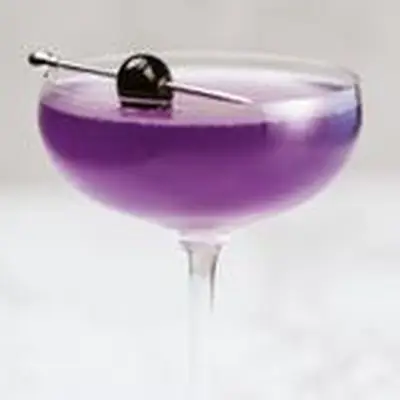 A vividly purple Aviation in a delicate coupe glass is garnished with a dark cherry, pierced on a silver pick