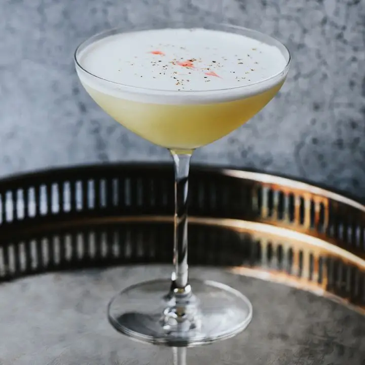 A curved Martini glass rests on a high-rimmed round bar tray. The drink within the glass is golden yellow with a layer of white foam dappled with nutmeg and bitters. The background is mottled gray. 