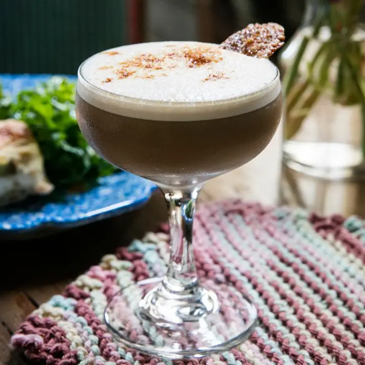 In a busy photo, a coupe glass sits on a colorfully knitted mat. The glass holds a dark brown cocktail with a foamy white head dotted with bitters and garnished with a thin cookies. A plate of food and a bottle of water are in the background.