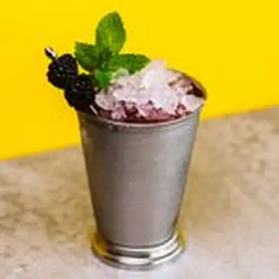 A silver julep cup is beaded with condensation. The cup holds a violet liquid, a pile of crushed ice, a sprig of mint, and two blackberries pierced on a silver pick. The drink sits on a tan surface with a bright yellow wall behind it. 