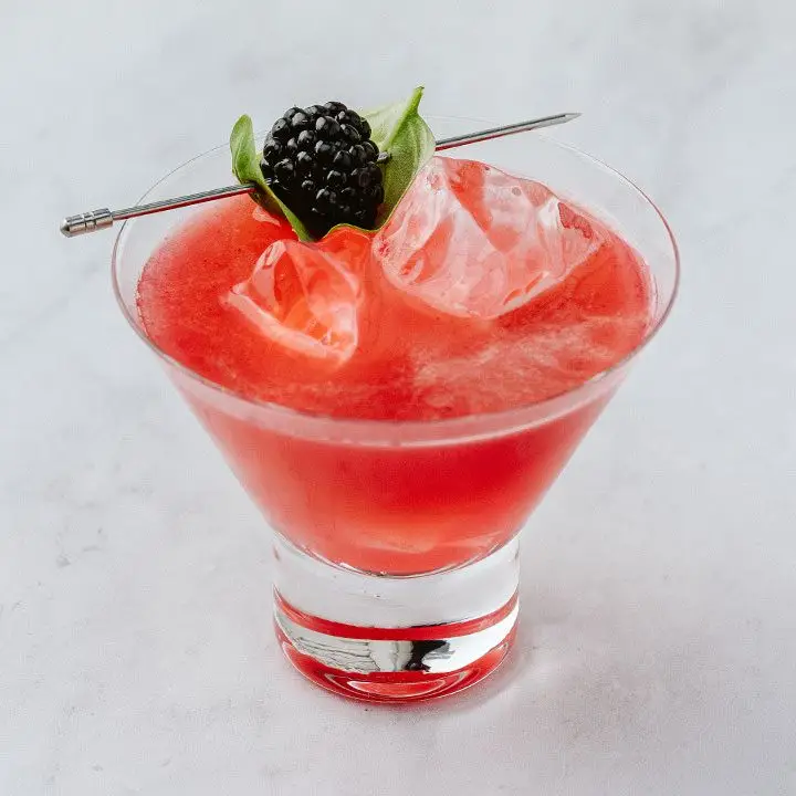 red-hued Black Widow cocktail in a stemless cocktail glass, garnished with skewered blackberry and basil leaf