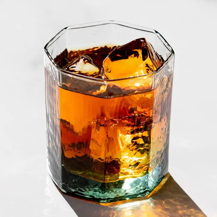 A hexagonal rocks glass sits on a dramatically lit white surface. It’s filled with ice cubes and an amber drink.