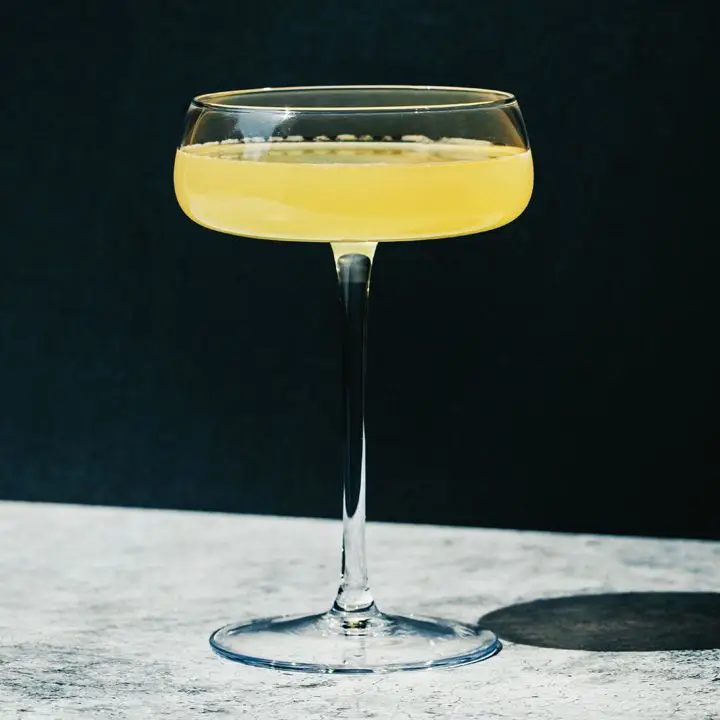 A wide, flat coupe rests on a marble surface. It’s filled with a golden cocktail.