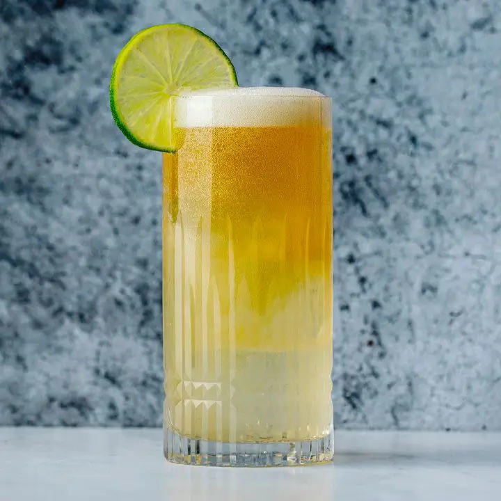 A highball glass is filled with a sparkly Margarita that gets darker towards the top of the drink, which also has a short white foam cap. It’s garnished with a lime wheel and set against a blue marble backdrop.