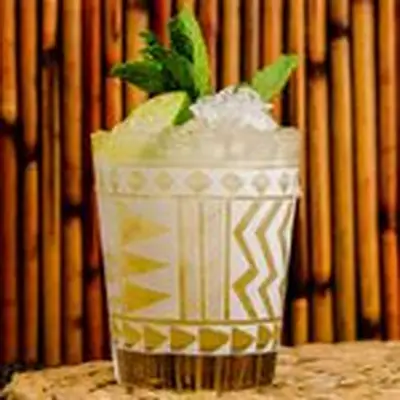A Tiki glass with white markings holds a golden Mai Tai. Crushed ice, mint and a lime wedge pick from above the lip of the glass. The background is wood lines, and the drink rests on a wicker mat.