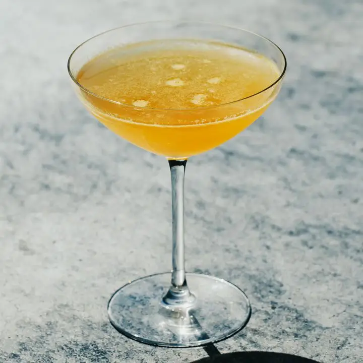A narrow-stemmed cocktail glass with a wide brim holds a pale orange cocktail. A few bubbles on the surface reveal how recently it was poured, and the orange of the drink is set off against the gray marble backdrop.