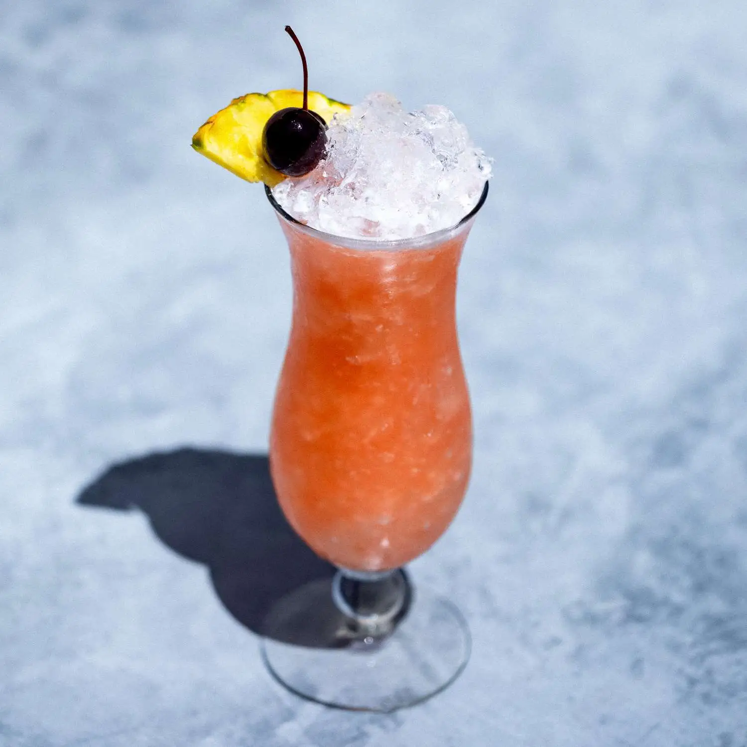 Rum Runner cocktail, orange-pink, in curved Hurricane glass over crushed ice, garnished with pineapple wedge and cherry