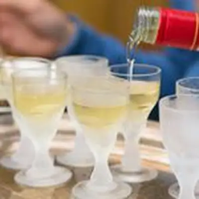 In the foreground are a number of frosty snifters, filled with a yellow-hued absinthe. More absinthe is flowing into one from a red-necked bottle. In the background, out of focus, is an arm in a blue shirt.