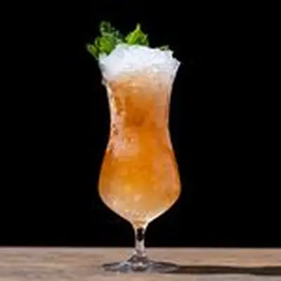 Zombie cocktail in curved Hurricane glass, orange hued over crushed ice with mint sprig garnish, on back background