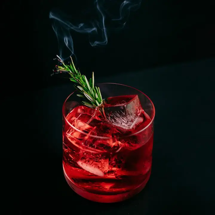 red-colored Rosemary Mezcal Negroni cocktail in a rocks glass, garnished with a lit rosemary sprig releasing a wisp of smoke