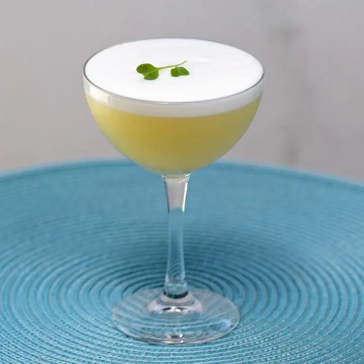 A rounded coupe sits on a sky blue placemat. The drink within is golden yellow, topped with thick white foam with a micro green floating on it.