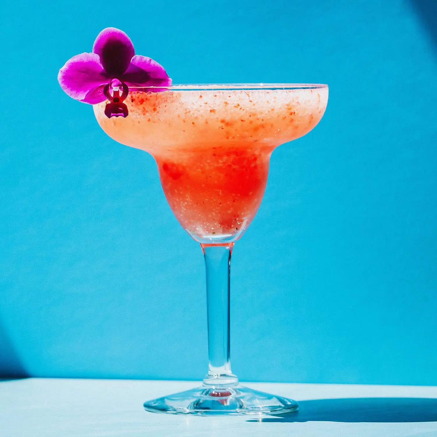 Frozen strawberry daiquiri in stemmed margarita glass, with purple orchid flower garnish, on blue background