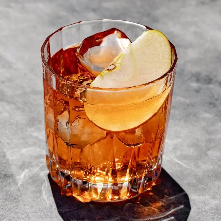 A beveled rocks glass with a reddish-brown stirred cocktail, three clear ice cubes, and a thin apple slice