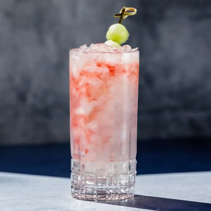 A Collins glass containing a translucent white cocktail with swirls of red raspberry liqueur, garnished with a skewered honeydew melon ball