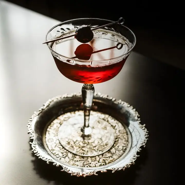 a la louisiane cocktail with cherry garnish served in a coupe glass on an ornate round metal coaster