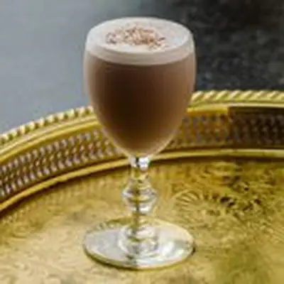 A port glass rests on a golden bar tray. The glass is filled with a dark brown drink with a fluffy white foam head, dusted with nutmeg.