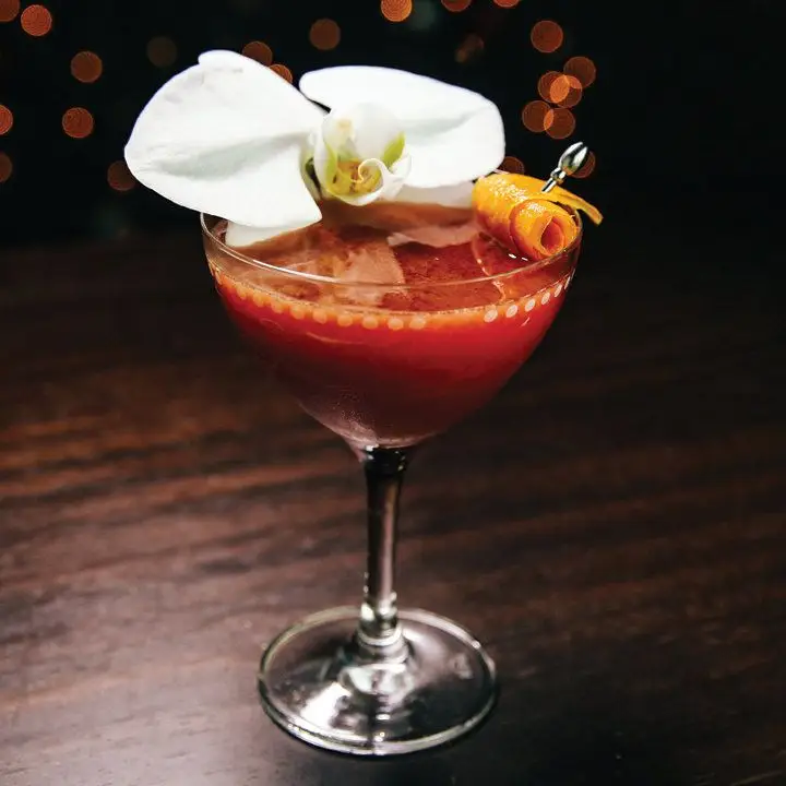 red-hued Robin Redbreast cocktail in a coupe, with a skewered orange twist and edible orchid