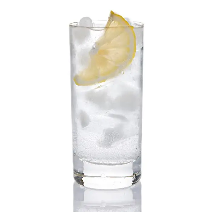 A Collins glass filled with ice, a vodka and aquavit Collins, and a lemon wedge is set against a stark white backdrop. 