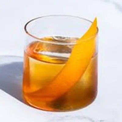 Old Fashioned cocktail in a rocks glass with single large ice cube and orange twist, on light marble background