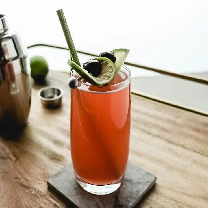 A highball glass rests on a square marble coaster on a wooden bar tray with copper handles. The glass is filled with an orange cocktail and is garnished with two lime wheels cradling a cherry each, all pierce on silver pick. A green straw extends from the glass, and a shaker and lime are behind it.