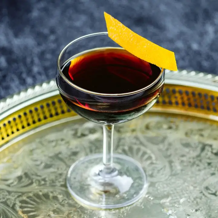 deep-brown Downhill Daring cocktail in a coupe glass with an orange peel on the rim, served on a round metal tray