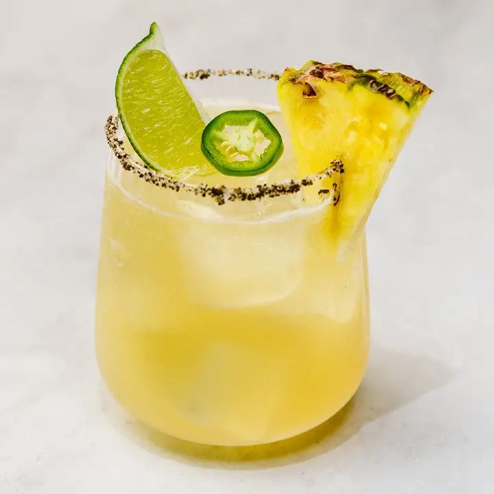 yellow-hued Pineapple Jalapeno Margarita in a sugar-and-black-pepper-rimmed rocks glass, garnished with pineapple, lime wedge and jalapeño slice