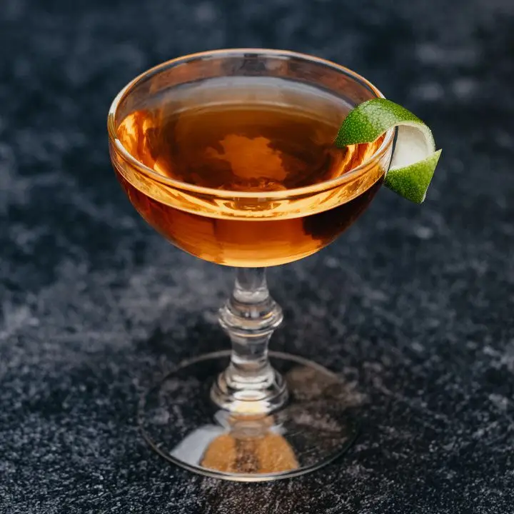 A knobbed cocktail couple rests on a dark marbled surface. The drink within is golden and garnished with a lime peel.